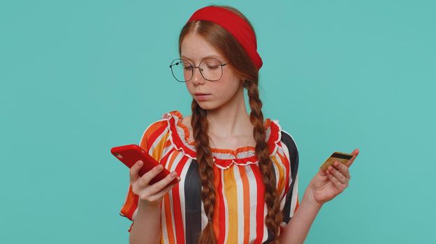 Sincere redhead girl customer using credit bank card and smartphone while transferring money, purchases online shopping, payment. Finance, internet. Young teenager child isolated on blue background