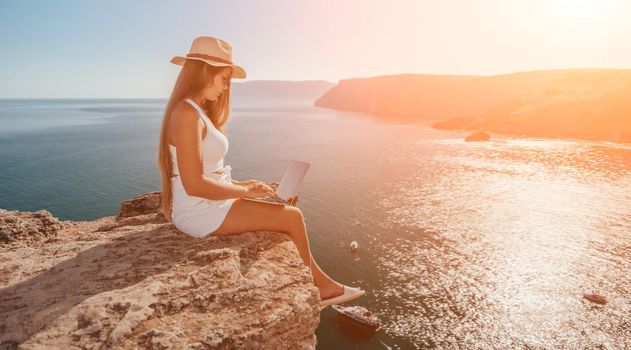 Successful business woman in yellow hat working on laptop by the sea. Pretty lady typing on computer at summer day outdoors. Freelance, travel and holidays concept.