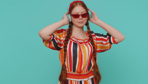 Portrait of redhead girl in dress listening music via headphones and dancing disco fooling around having fun expressive gesticulating hands. Young teenager child on blue studio wall background