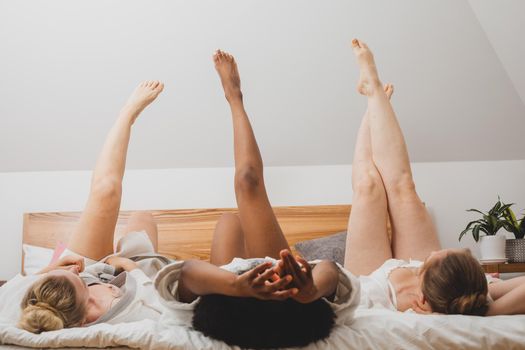 Three young women in a bathrobes are lying on the bed with their legs raised up