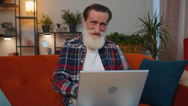 Overjoyed senior old grandfather on home couch with laptop scream in delight raise hands in triumph winner celebrate success win money in lottery. Excited elderly retired man get online good news