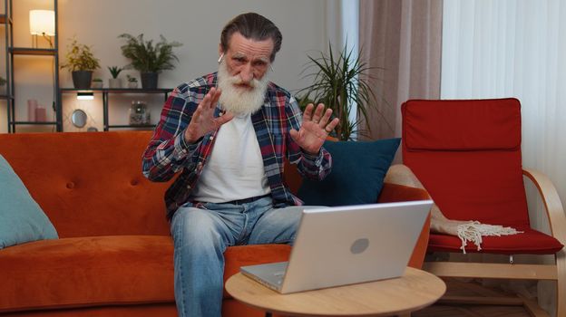 Senior old grandfather sitting on couch, looking at laptop, making video webcam conference call with friends or family, enjoying pleasant conversation. Elderly man laughing, waving hello alone in home