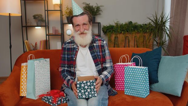 Happy senior old grandfather wears festive birthday cap hat hold gift box with ribbon congratulating. Man celebrating anniversary party event opening delivery greeting present on couch at home indoors