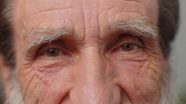 Extreme close-up macro portrait of wrinkled bearded face. Old senior beautiful man's eyes, smiling looking at camera. Brown eyes of elderly guy male model. Mature grandfather opening wide his eyes