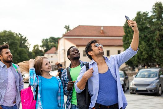 The group of adult multiethnic students take a selfie using a smartphone. The students with backpacks outdoors is smiling while looking at the phone