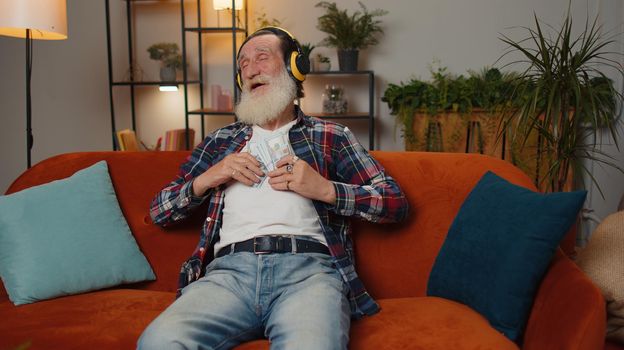 Successful rich senior businessman grandfather man holding money cash cheering up with high profits, lottery game win, satisfied of wealth income earnings salary on home couch, dancing listening music