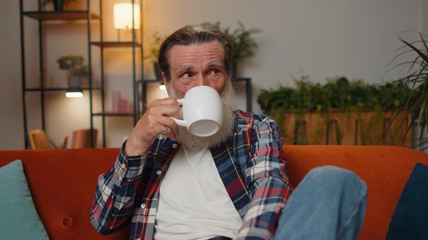 Attractive smiling senior old grandfather man drinking a cup of warm coffee or herbal tea sitting at home living room couch in the morning. Elderly retired grandpa pensioner enjoying comfortable relax