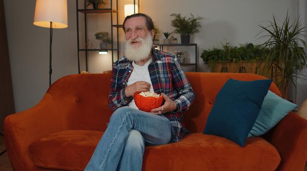 Excited elderly grandfather sitting on sofa eating popcorn and watching interesting tv serial, sport game film, online social media movie content online at home. Man enjoying domestic entertainment