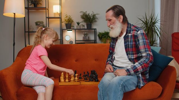 Smiling senior old grandfather man playing chess with teen granddaughter at home living room. Retired elderly grandparent enjoying play board game, resting with child girl kid. Family leisure activity