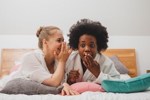 Two young women of different nationalities are lying on the bed at the spa and talking. The african woman is telling secret to her friend