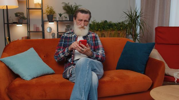 Senior old grandfather sitting on sofa uses mobile phone smile at modern home apartment. Elderly man texting share messages content on smartphone social media applications online, watching relax movie