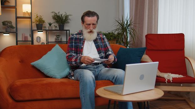 Stressed senior old grandfather man at home room looking at unpaid bank debt bills, doing paperwork, planning budget, calculate finances mortgage payments. Elderly retired pensioner