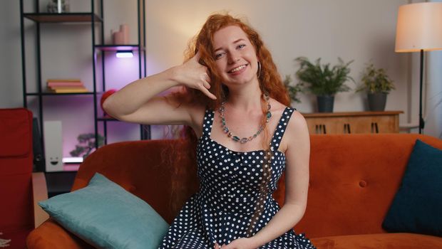 Call me, here is contact number. Redhead young woman looking at camera advertising doing phone gesture like says hey you call me back at home apartment. Ginger cute girl in living room sitting on sofa