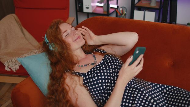 Happy overjoyed redhead young girl in wireless headphones relaxing lying on sofa at home choosing listening favorite energetic disco rock n roll dancing music in smartphone. People weekends activities