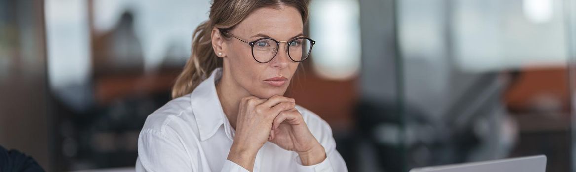 Serious businesswoman with glasses working on laptop at her workplace in modern office.