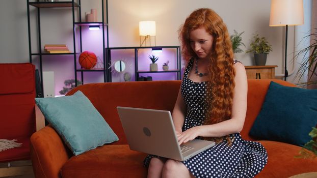 Redhead young woman in polka dots dress freelancer in home living room sits on couch, opens laptop start working. Ginger girl works on notebook, sends messages, makes online purchases, watching movies