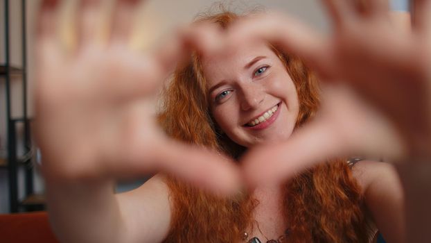 I love you. Redhead young woman makes symbol of love, showing heart sign to camera, express romantic feelings, express sincere feelings. Charity, gratitude. Girl close-up face with freckles indoors
