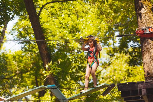 The concentrated little girl carefully steps on the suspension bridge. The girl in protective gear is playing in the rope park