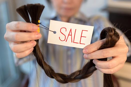 Long brown cut braid with tag in female hands, sale of woman cut hair, depth of field, front selective focus
