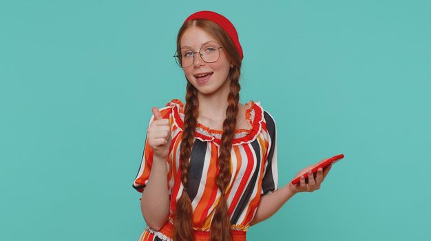 Beautiful red hair girl using mobile phone typing new post on web sms message, browsing, searching addiction of social networks. Young ginger teenager child studio shot isolated on blue background