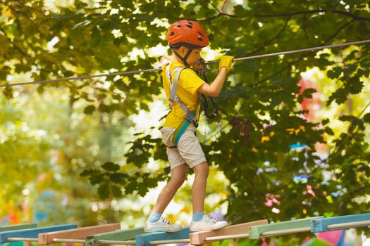 Portrait of a concentrated boy who is climbing in the rope park in summer. He is wearing a protective helmet and gloves