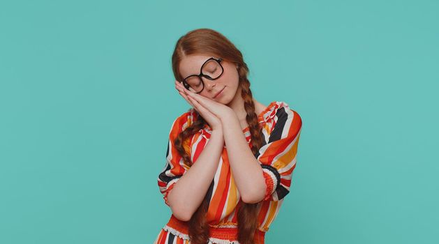 Tired redhead girl going to sleep laying down on his palms, yawning, dozing off during day, taking nap and resting, comfortable bedtime. Teenager ginger child kid isolated on blue studio background