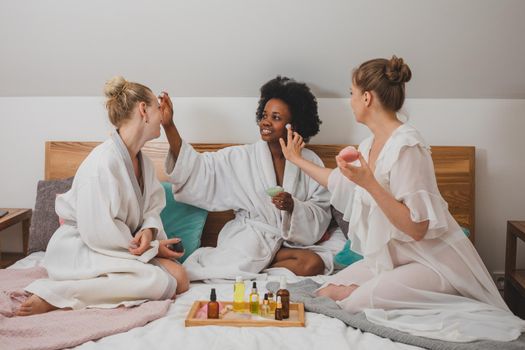 Three smiling girlfriends sit on the bed and smear each other with cream. The young women in bathrobes have a spa party