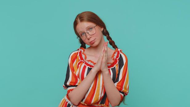 Please, God help me. Redhead girl praying, looking upward and making wish, asking with hopeful imploring expression, begging apology. Young teenager ginger child kid isolated on blue studio background