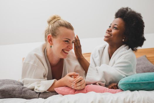 Two young women of different nationalities are lying on the bed at the spa and talking. The african woman is telling secret to her friend
