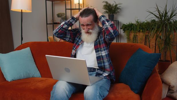 Sad senior old grandfather man use laptop typing browsing working, loses becoming surprised sudden lottery results, bad news, fortune loss, fail. Elderly retired pensioner at home living room couch