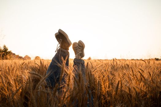 Backlit light picture couple's legs in sneakers over grain field and sunset sky. Man and woman laying among spikelets, holding legs up.
