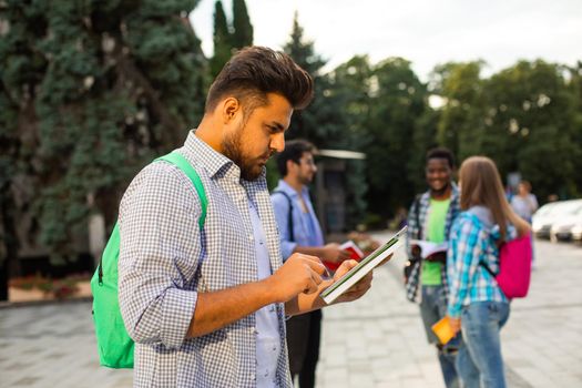 The young indian student wearing shirt with a backpack is holding a tablet and book in his hand.