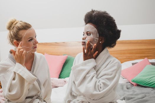 Two young women in masks are sitting next to a set for spa treatments. The women dressed in bathrobes are sitting on the bed and look at the camera