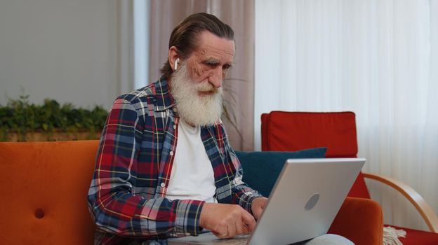 Senior old grandfather man freelancer at home room couch working on laptop. Elderly retired grandpa works on notebook, sends messages, makes online order purchases or food delivery, watching movies