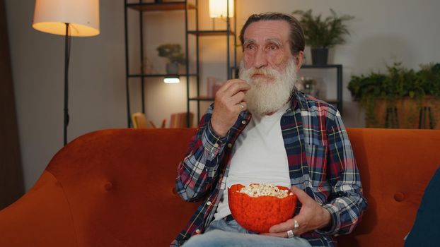 Excited mature old grandfather sitting on sofa eating popcorn and watching interesting tv serial, sport game film, online social media movie content online at home. Man enjoying domestic entertainment