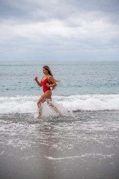 A beautiful and sexy brunette in a red swimsuit on a pebble beach, Running along the shore in the foam of the waves.