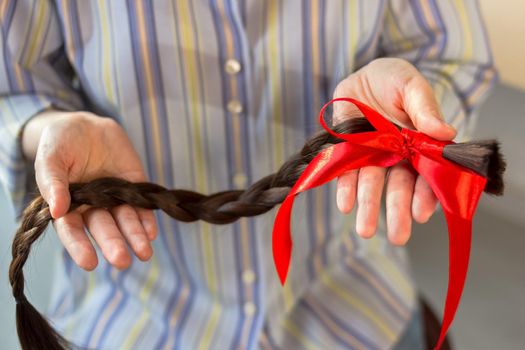 Long brown cut braid with red ribbon bow in female hands, woman donation of haircut, faceless concept