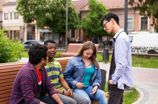 Group of cheerful diverse friends meeting outdoors at the campus park, sitting on wooden bench. Side view lovely girl in denim shirt having conversation with multiracial male friends.