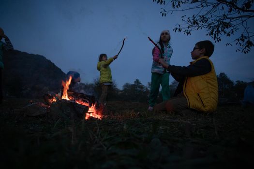 Children after hiking have a picnic - group of happy friends frying sausages on campfire
