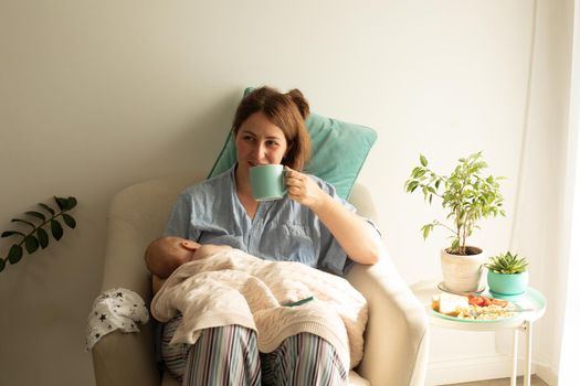 Woman drink tea or coffee during process of breastfeeding of newborn baby. Comfortable breastfeeding