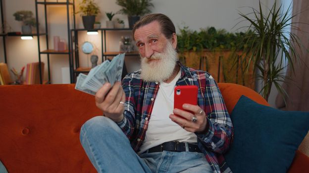 Planning family budget. Smiling senior old grandfather counting money cash, use smartphone calculate domestic bills at home. Joyful man satisfied of income and saves money for planned vacation, gifts
