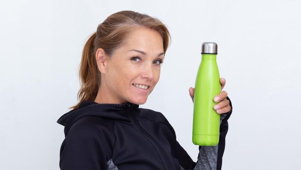 Caucasian smiling middle aged woman in sportiv dress holding light green thermos bottle with water on white background looking at camera