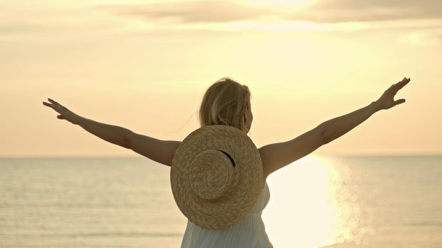 Woman in white dress and straw hat standing with open arms on sea beach. Beauty, nature, travel, hope, harmony concept. High quality photo