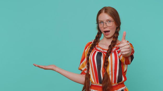Beautiful redhead girl showing thumb up, pointing empty place, advertising area for commercial text, copy space for goods promotion. Young ginger teenager child. Studio shot indoors on blue background