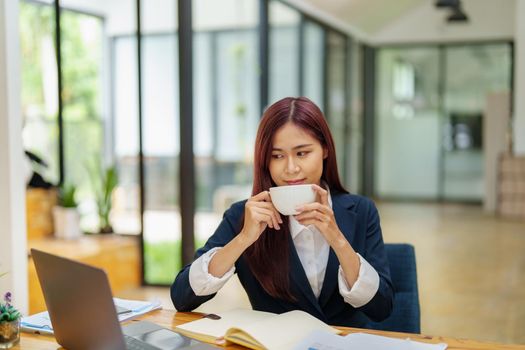 Asian female worker using computer and budget documents on desk.