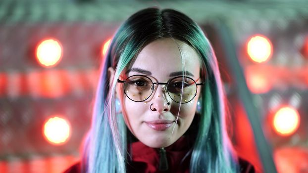 Hipster girl with blue dyed hair, golden sequins as freckles. Sad woman with nose piercing, transparent glasses, ears tunnels, unusual hairstyle stands in amusement night park. High quality photo