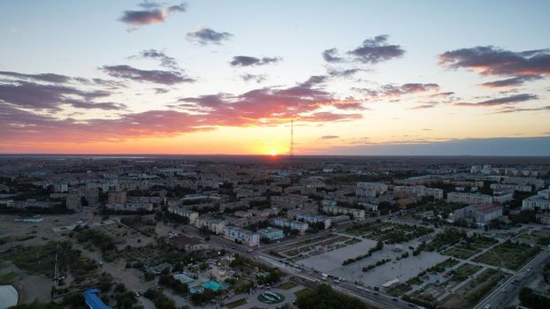 Orange sunset with a view of the city and the TV tower. The beautiful gradient of the sky and clouds shimmers from dark blue to light orange. Low houses, a green park and cars on the roads. Drone view