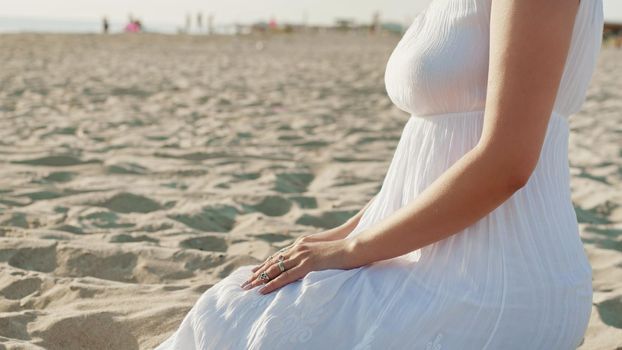 Woman in white dress and boho rings on hands sits on sandy beach alone. Meditation, immersion in inner world, unity with nature. High quality photo