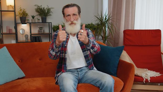 Great job. Senior old grandfather man showing thumbs up and nodding in approval, successful good work, satisfaction results at modern home apartment. Elderly retired pensioner in room sitting on sofa
