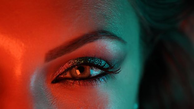 Close-up of woman eye under orange blue neon light. Female with beautiful makeup, glitter shadows. Beauty, cosmetics, night club lifestyle concept. High quality photo